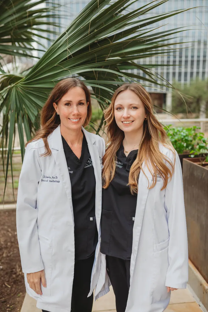Dr. Jill Davis and Dr. Margaret Thompson of Victory Hearing & Balance in white coats, standing outdoors with palm fronds and an Austin skyline in the background.